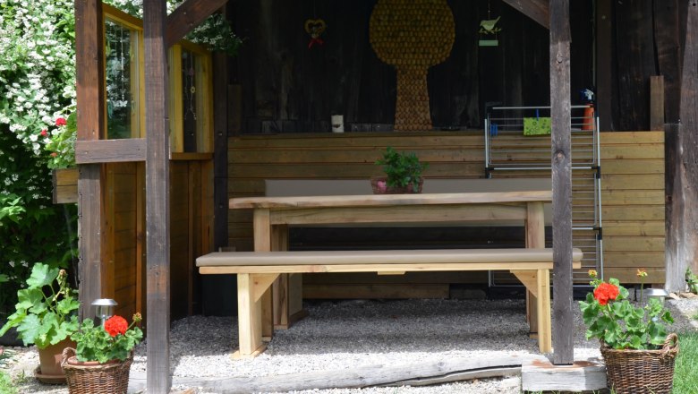 A rustic gazebo with a wooden table and benches, surrounded by plants and flowers in baskets.