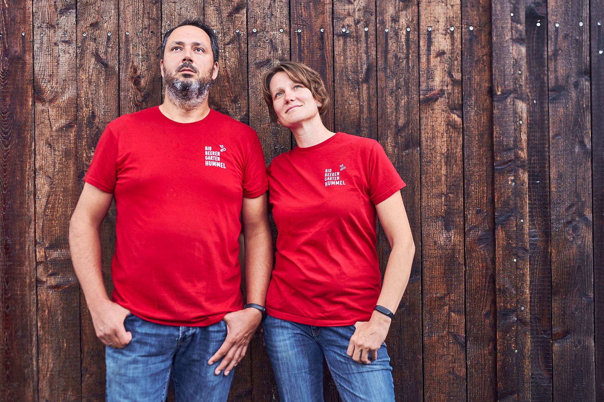 Two people in red T-shirts in front of a wooden wall.