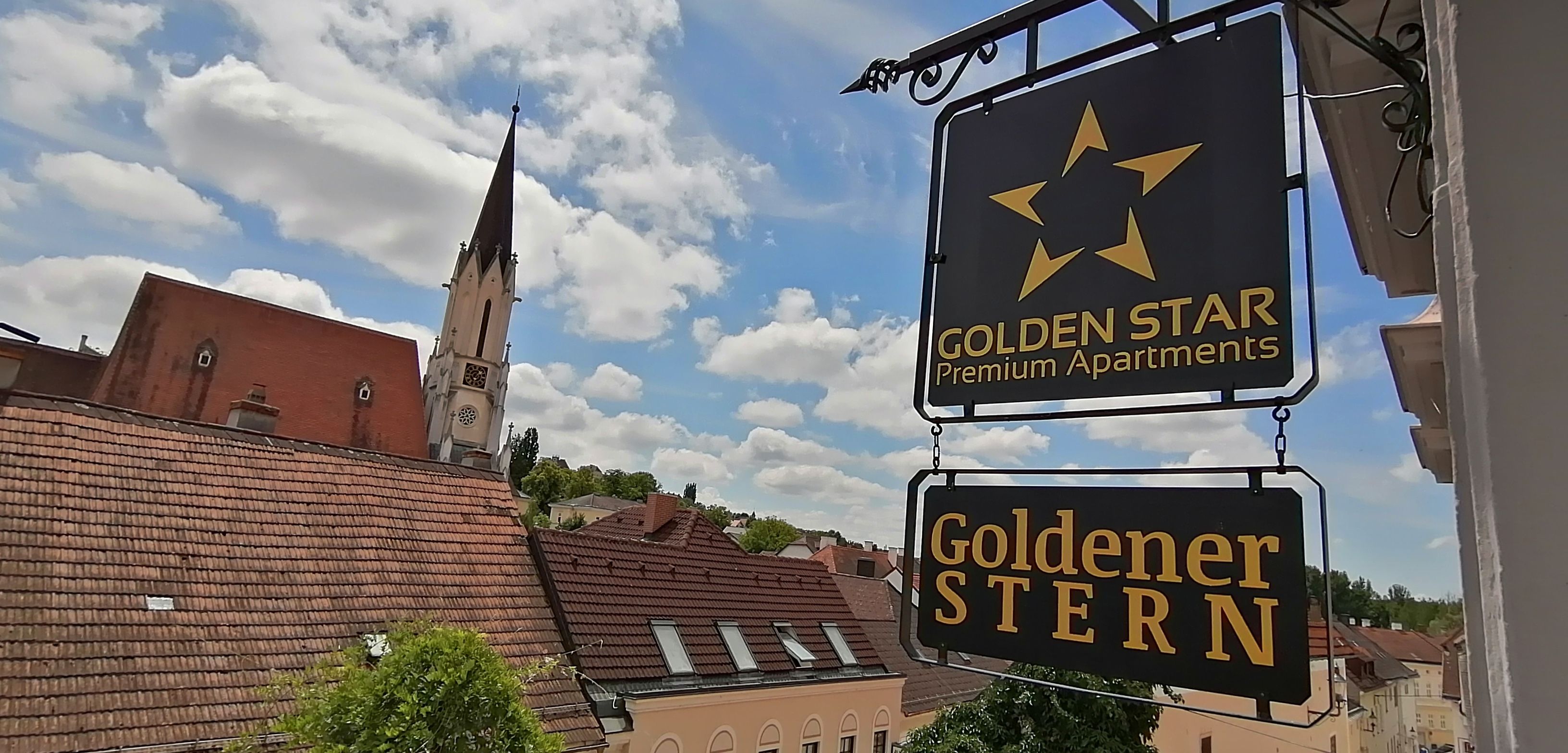 View of a sign with the inscription 'Golden Star Premium Apartments' and 'Golden Star' in front of a church tower and rooftops.
