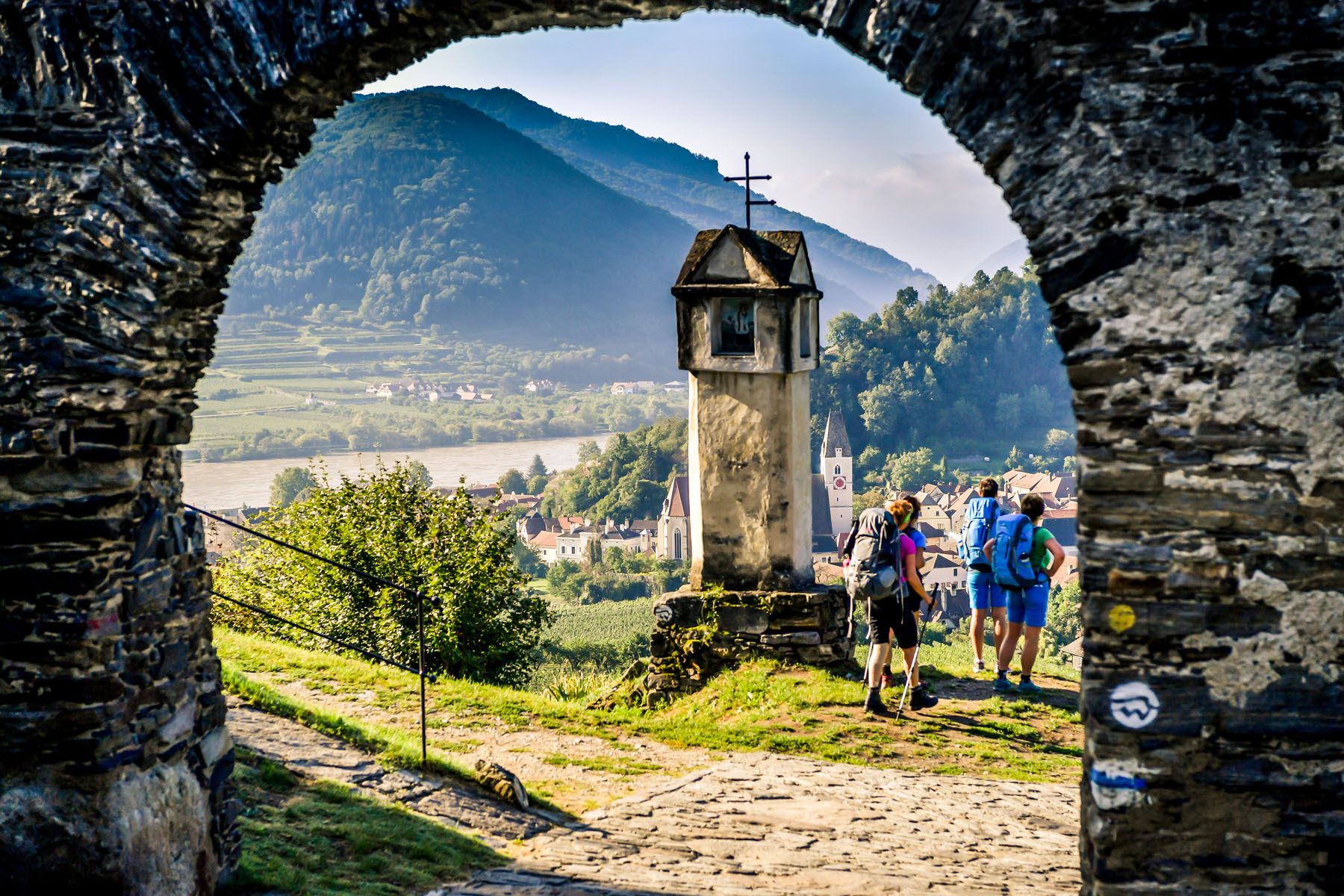 View through a stone gate onto a landscape with hills, a river and hikers.
