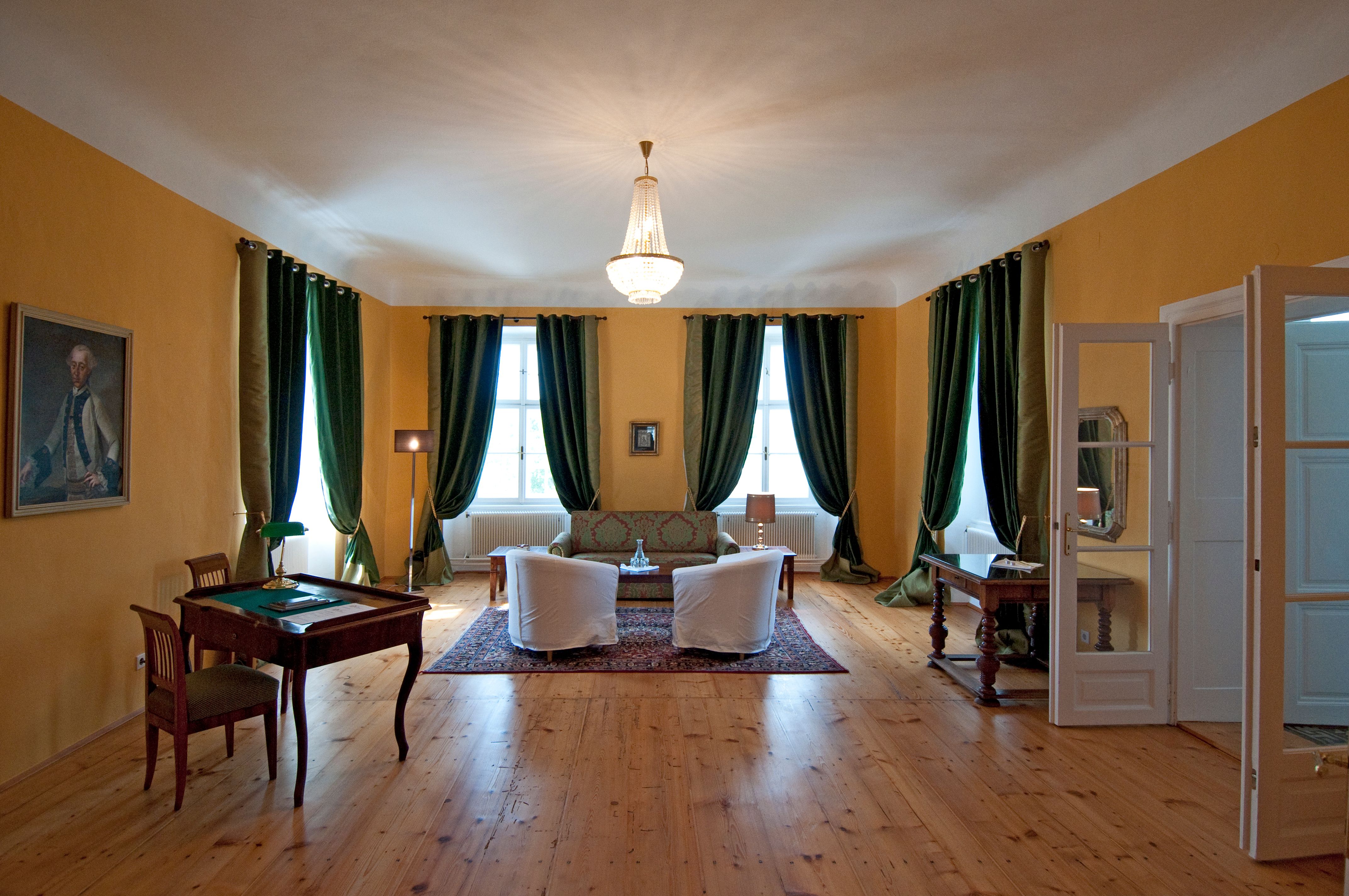 Elegant room with wooden floor, green curtains, two armchairs, a sofa and a desk. A chandelier hangs from the ceiling.