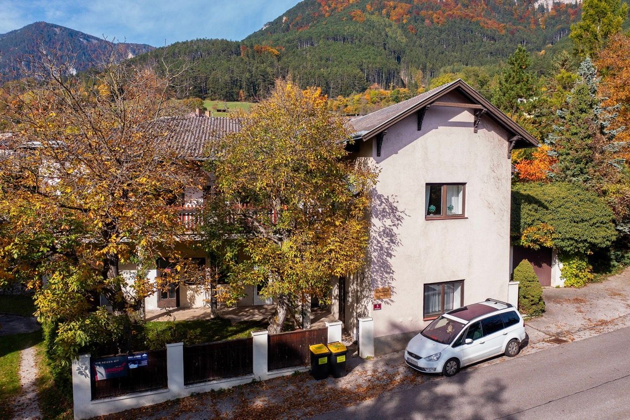 A two-story building with autumn trees and a parked car in front of it, surrounded by wooded hills.