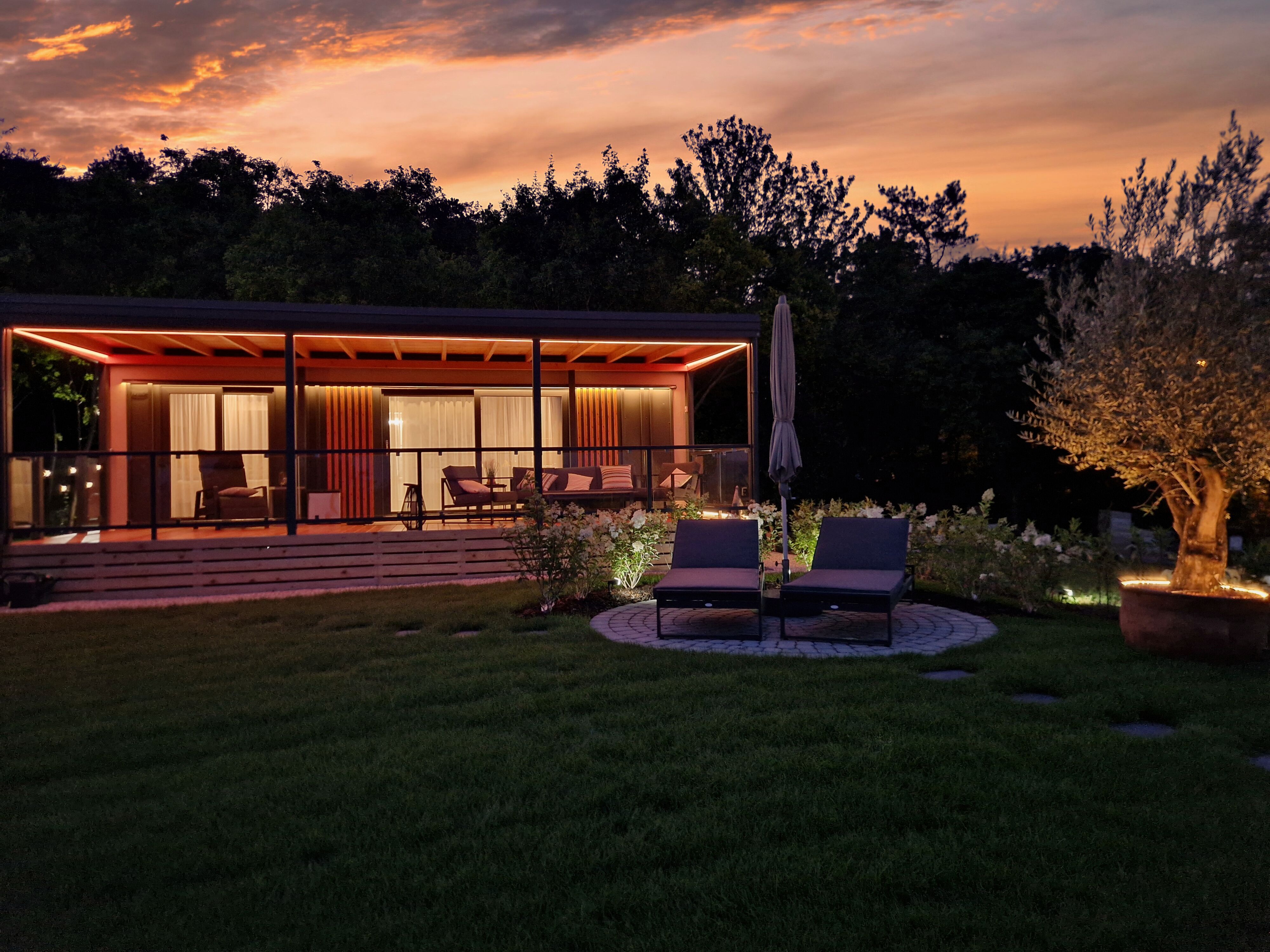 A modern chalet with terrace and sun loungers at sunset, surrounded by trees and an illuminated tree in the garden.