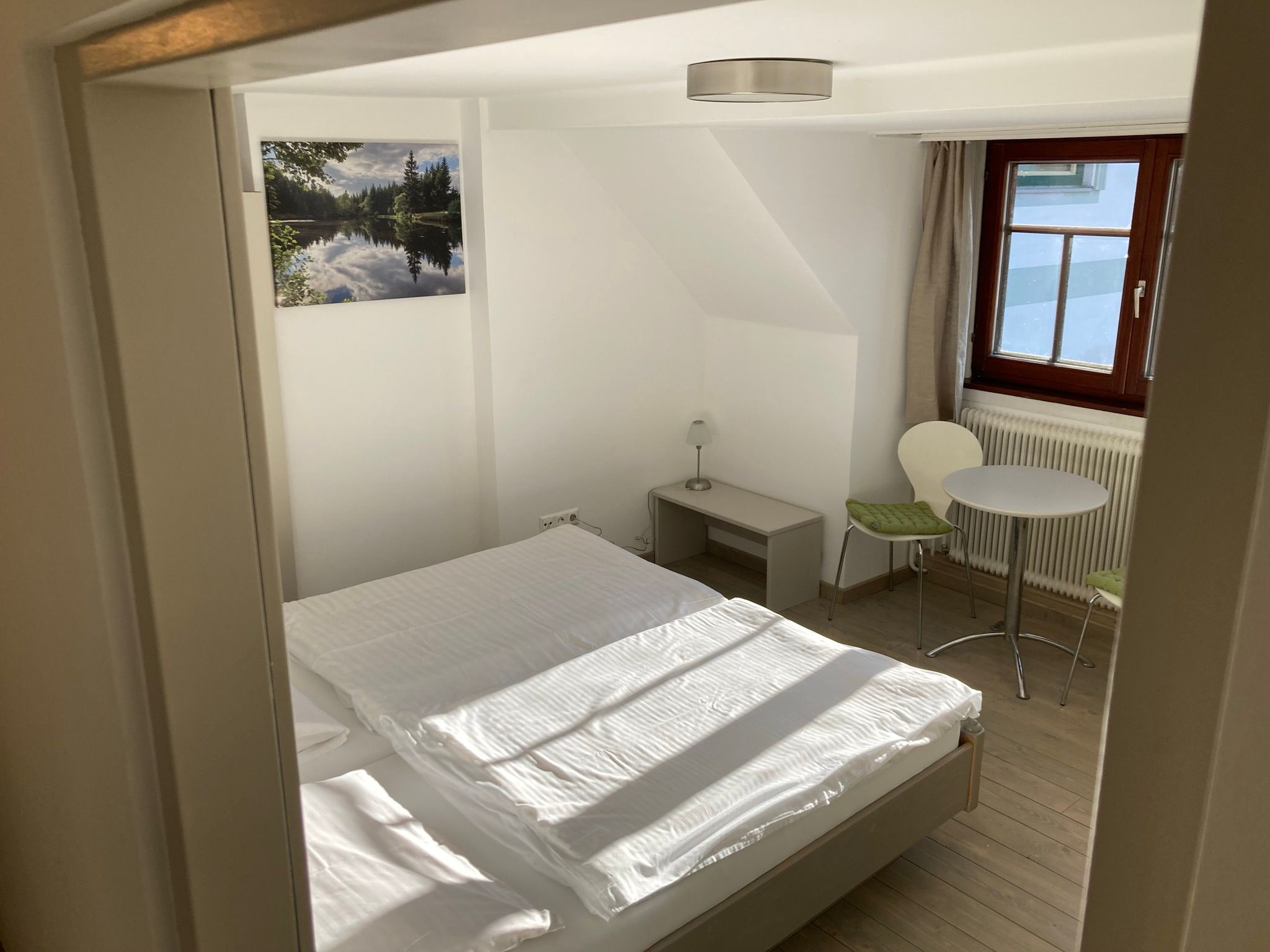 A simply furnished room with a bed, a small table and two chairs. A window lets in daylight and a picture hangs on the wall.