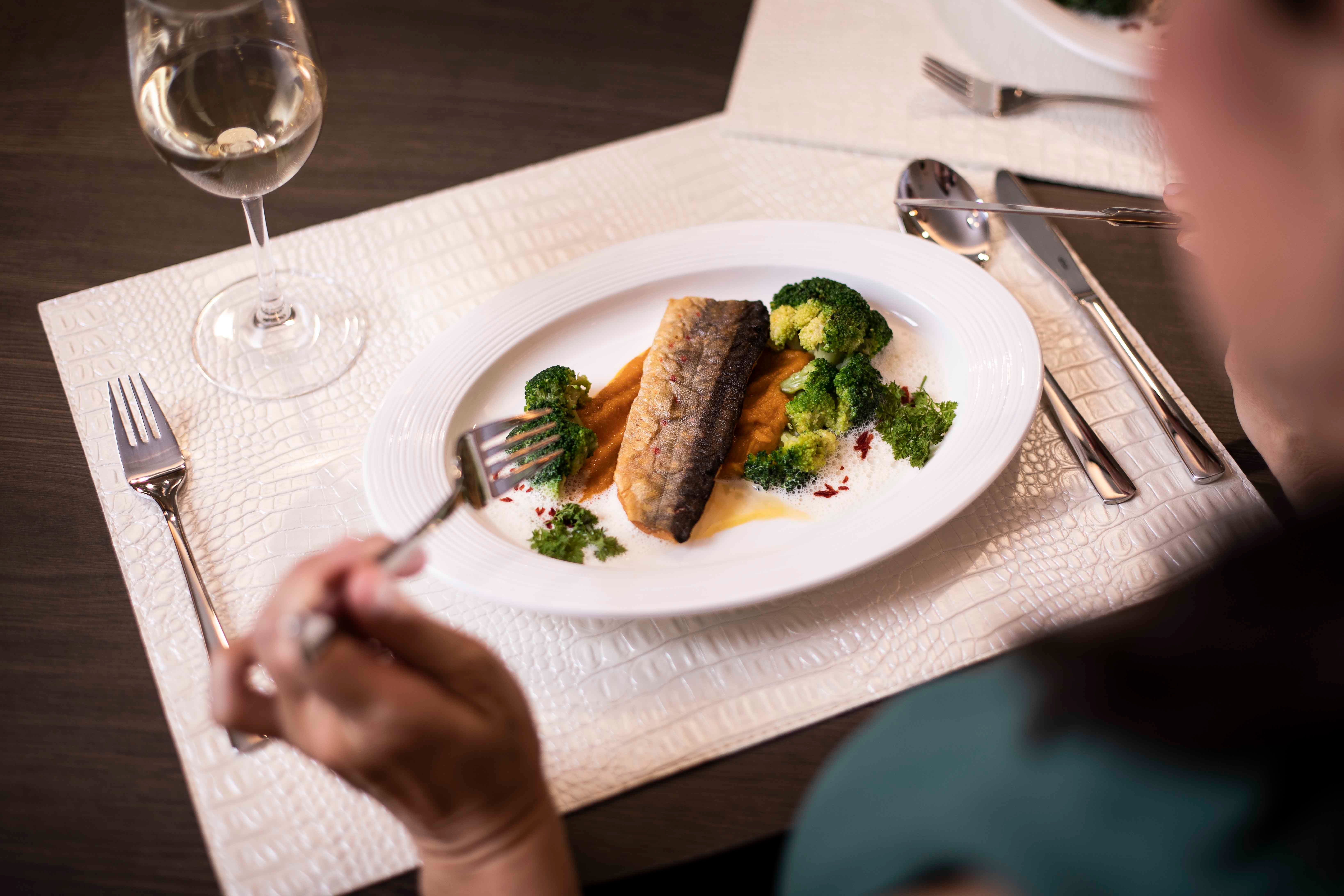 A plate with fish fillet, broccoli and sauce, with a glass of white wine next to it.