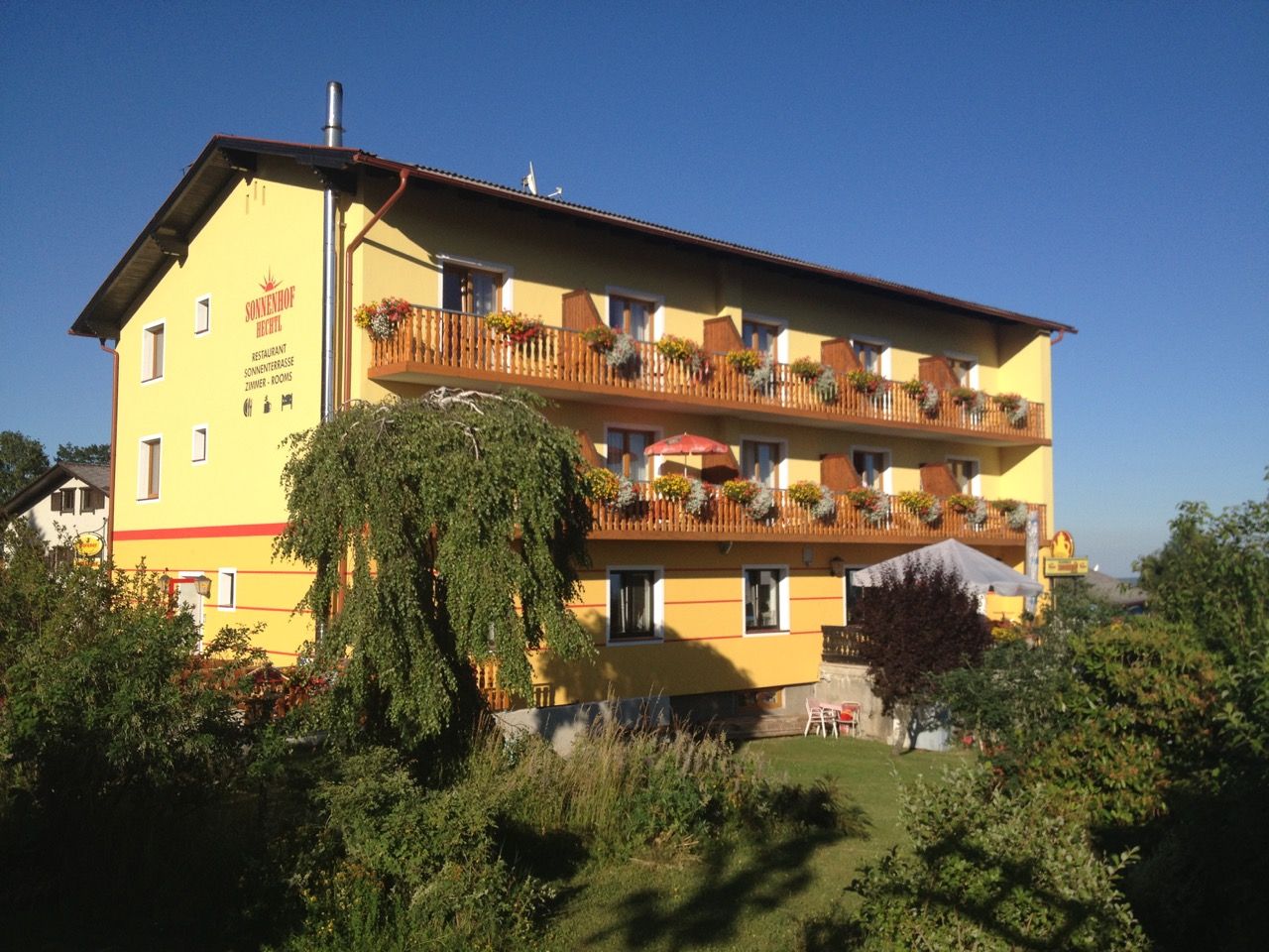 Yellow building with wooden balconies and flowers, surrounded by trees and garden, under a blue sky.