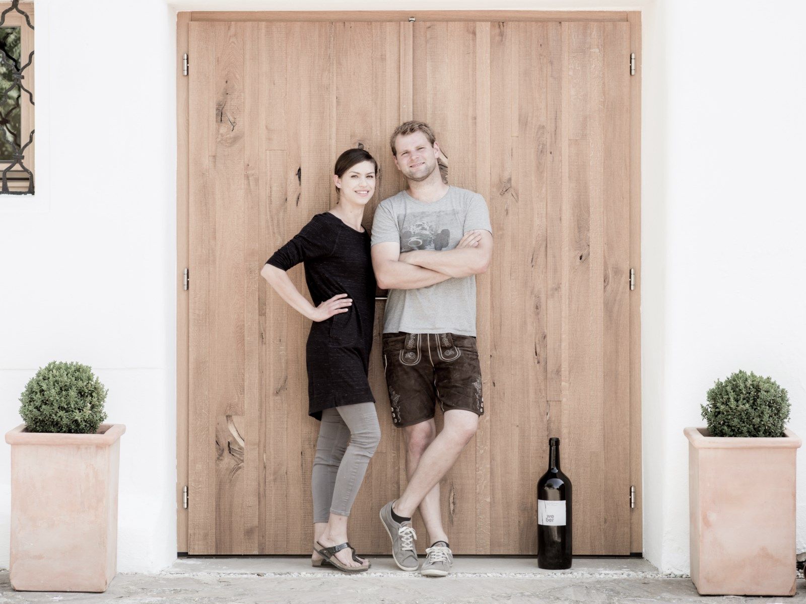 Two people are standing in front of a wooden door, next to a large bottle of wine and plants in pots.