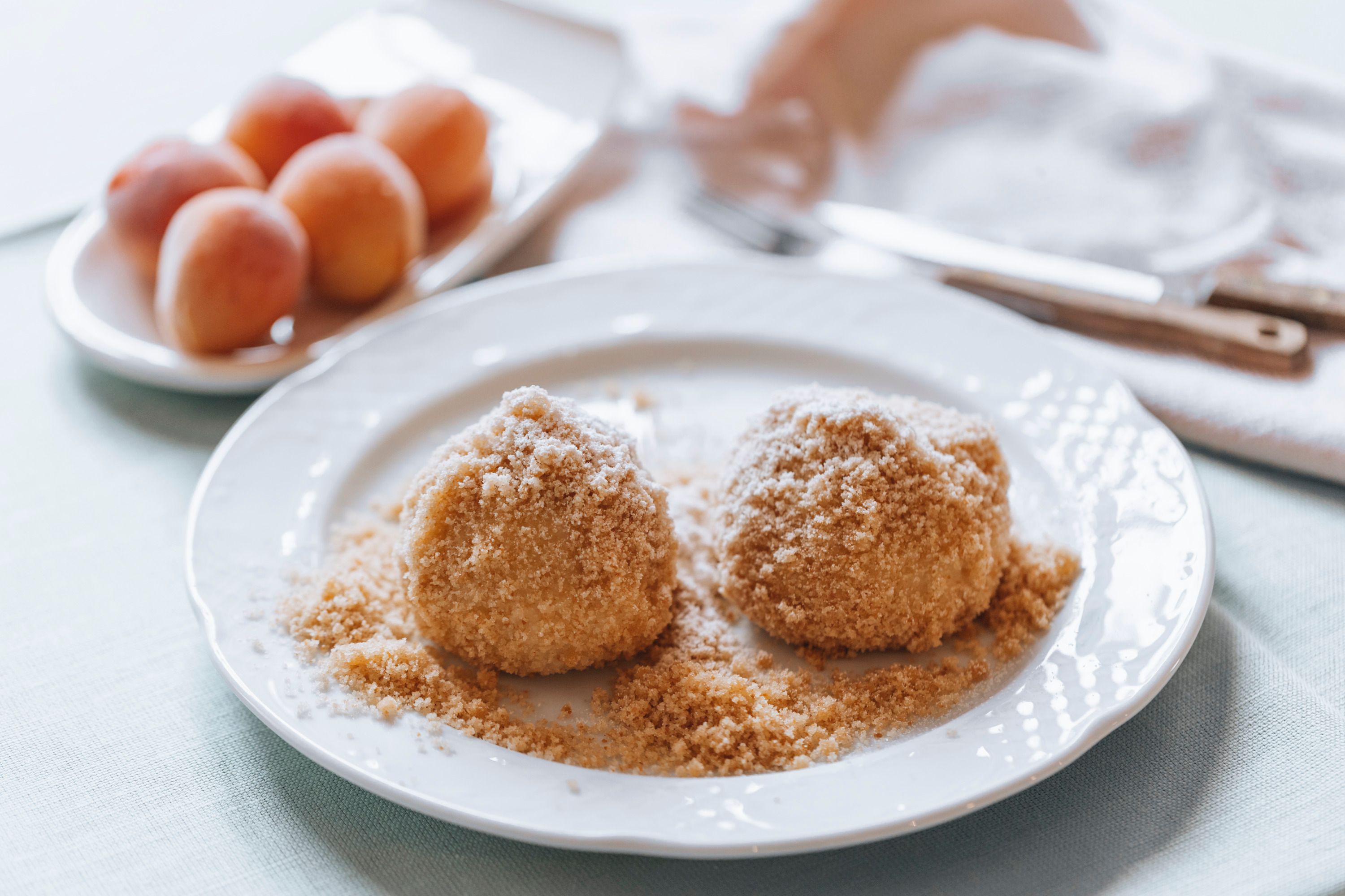 Two apricot dumplings on a plate with breadcrumbs, apricots in the background.