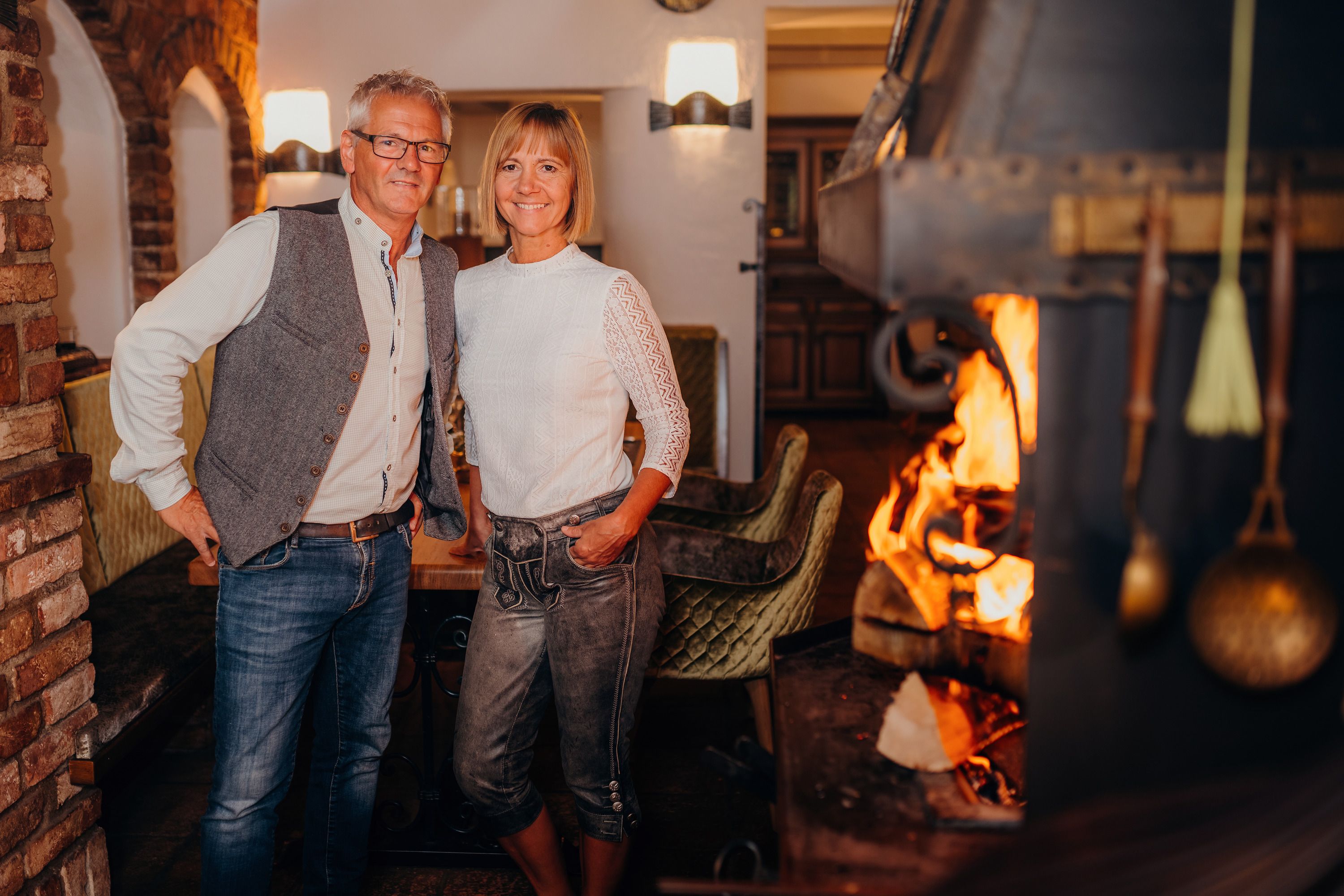 A man and a woman stand smiling in a cozy room with a fireplace.