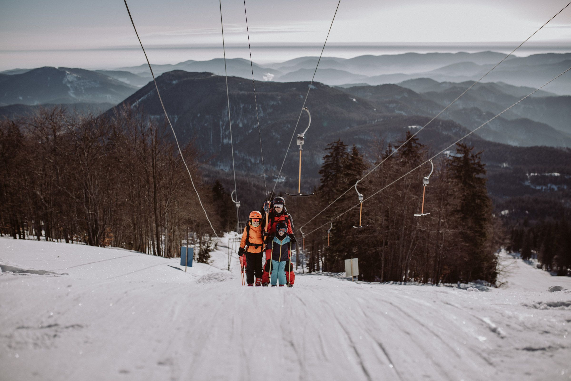 Three skiers on a drag lift in the Unterberg ski area, surrounded by snow-covered mountains and trees.