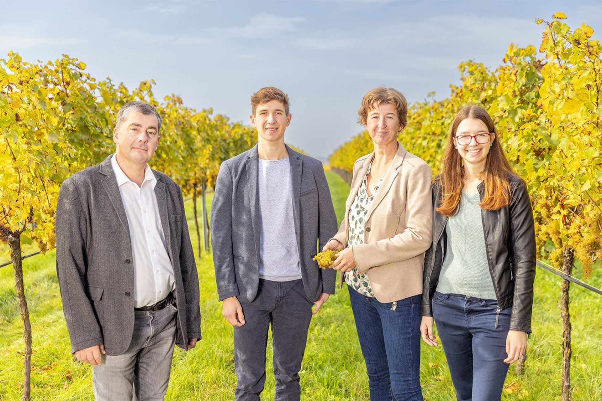 Four people stand together in the vineyard between the rows of vines; a natural, relaxed atmosphere with a rural background.