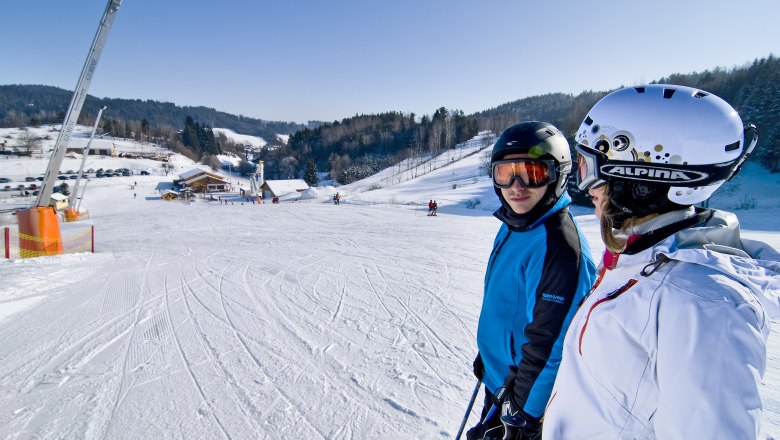 Two skiers stand on a ski slope in a snowy village with hills and trees in the background.