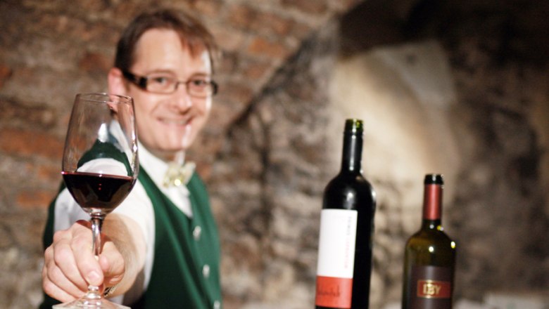 Teufl Inn, © weinfranz.at A man in traditional dress offers a glass of red wine, with two bottles of wine in the background.