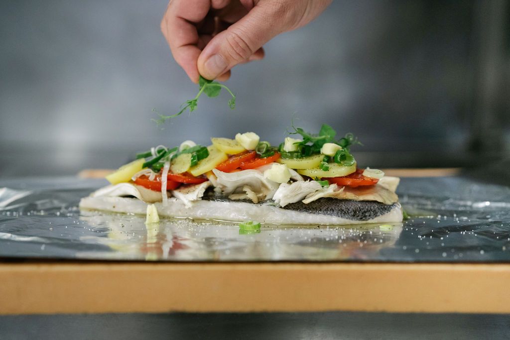 A fish fillet with vegetables and herbs on aluminum foil, a hand adds garnish.