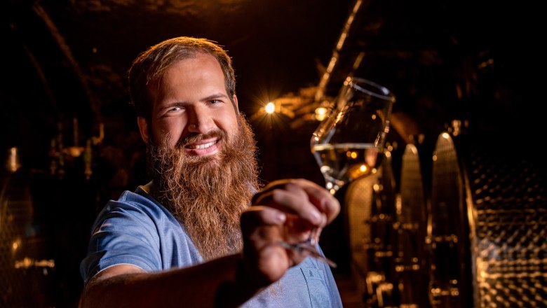 Man with beard holding a wine glass in a wine cellar.