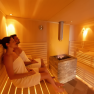 Two people relax in a sauna with wooden benches and a stove in the middle.
