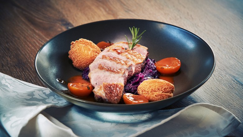 Gourmet dish with duck breast, red cabbage, potato dumplings and apricots on a black plate.