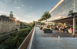 Panorama of a river landscape with a castle and modern architecture, surrounded by green hills.