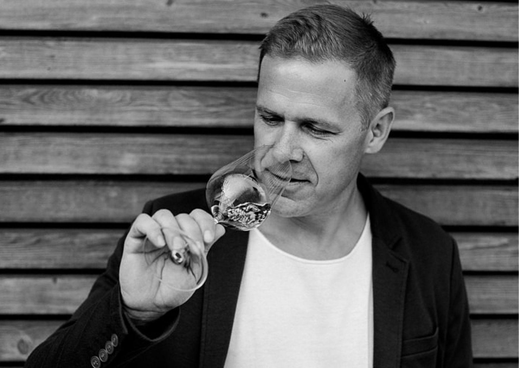 A man smells a glass of wine in front of a wooden wall.