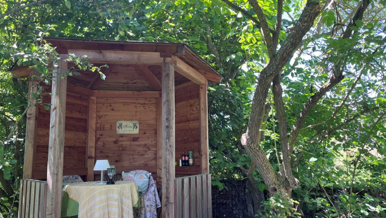 A cozy wooden pavilion in the garden, surrounded by trees and plants, with a table and chairs underneath.