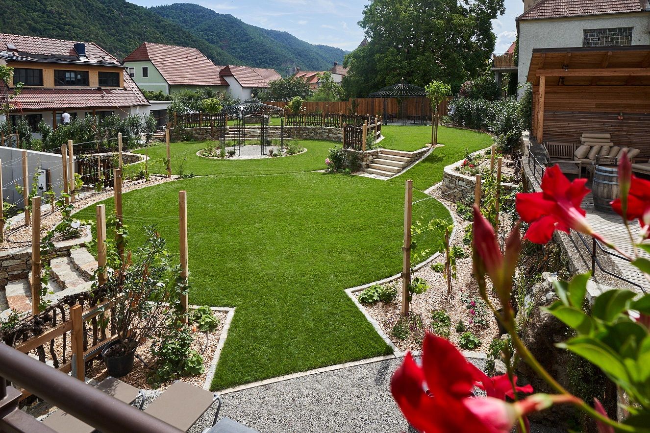 A well-tended hotel garden with a green lawn, flower beds and a pavilion, surrounded by houses and hills in the background.