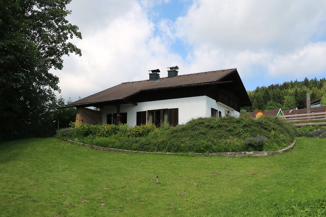 A white house with a brown roof in a green garden with trees and flowers.