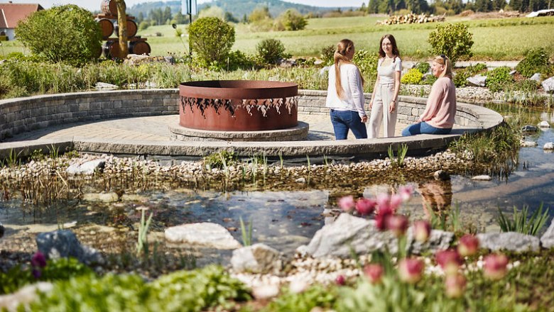 Three women sit in a garden with a small pond and a metal sculpture in the middle.