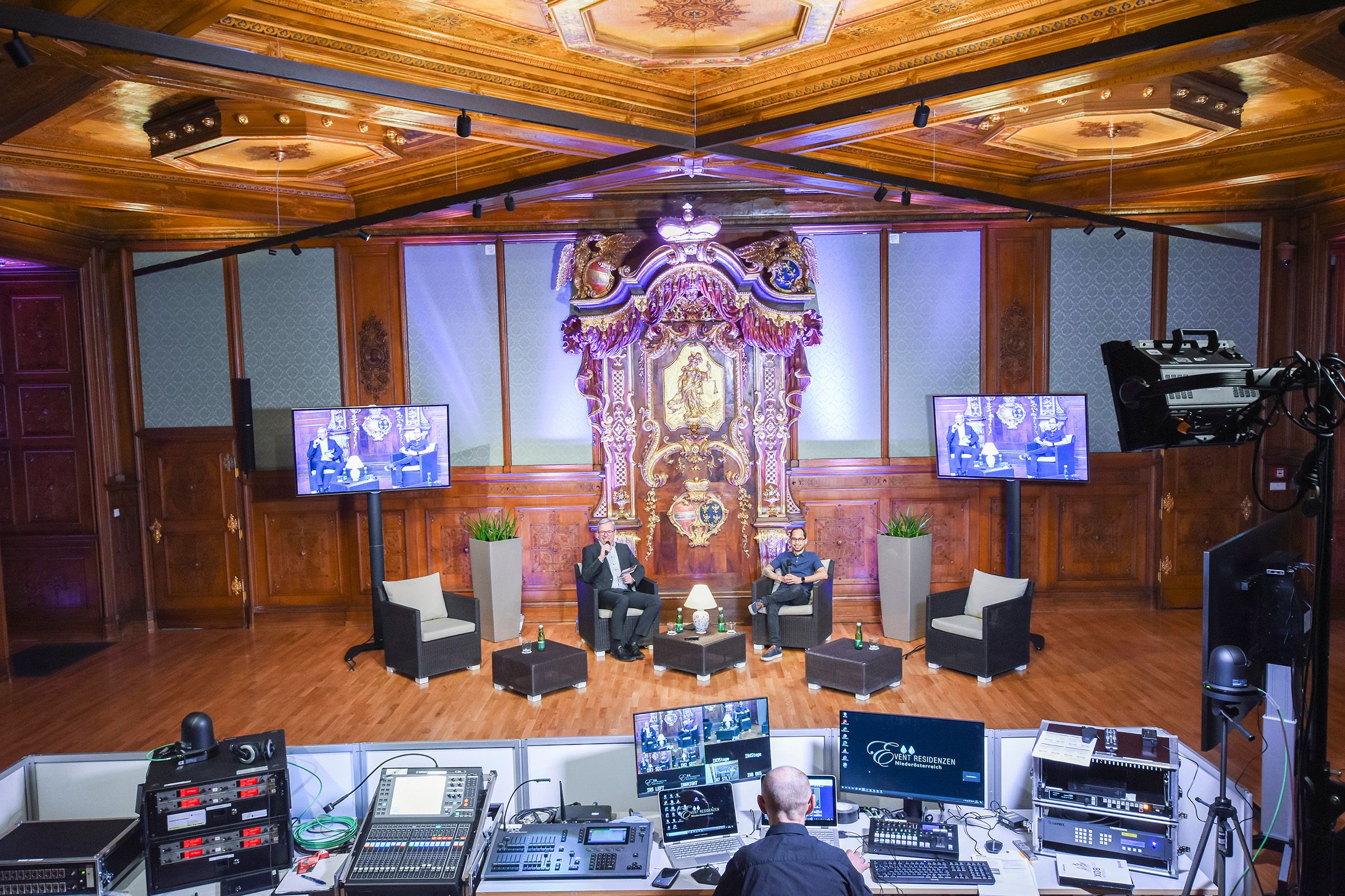 An opulent room with wood paneling and an elaborate altar. Two people sit on a stage, surrounded by technology and screens.