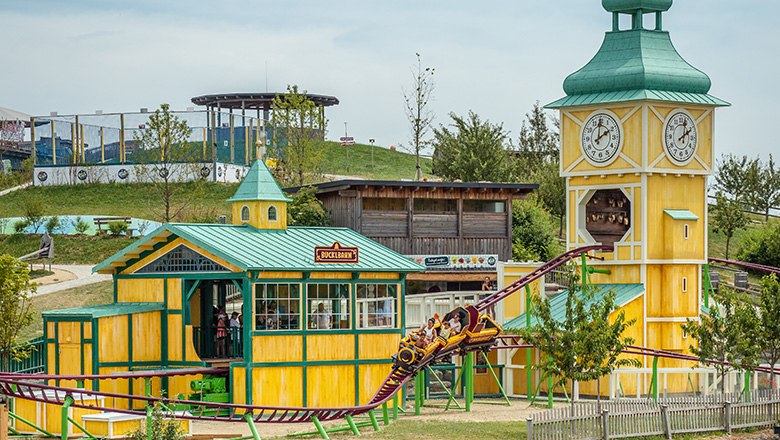 The "Bucklbahn" roller coaster in the Eis-Greissler Adventure Park with yellow buildings and a green hill in the background.