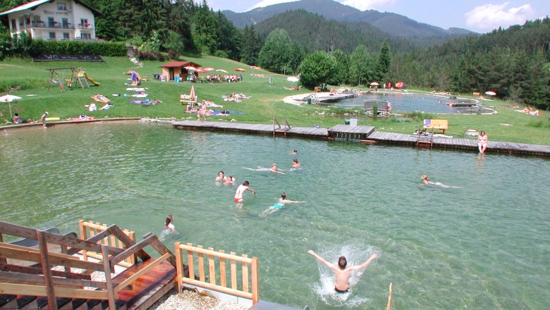 People swimming in a natural swimming lake with surrounding meadow and forest.