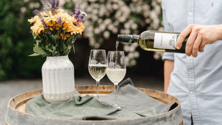 A person is pouring white wine into two glasses on a wine barrel, with a vase of flowers next to it.