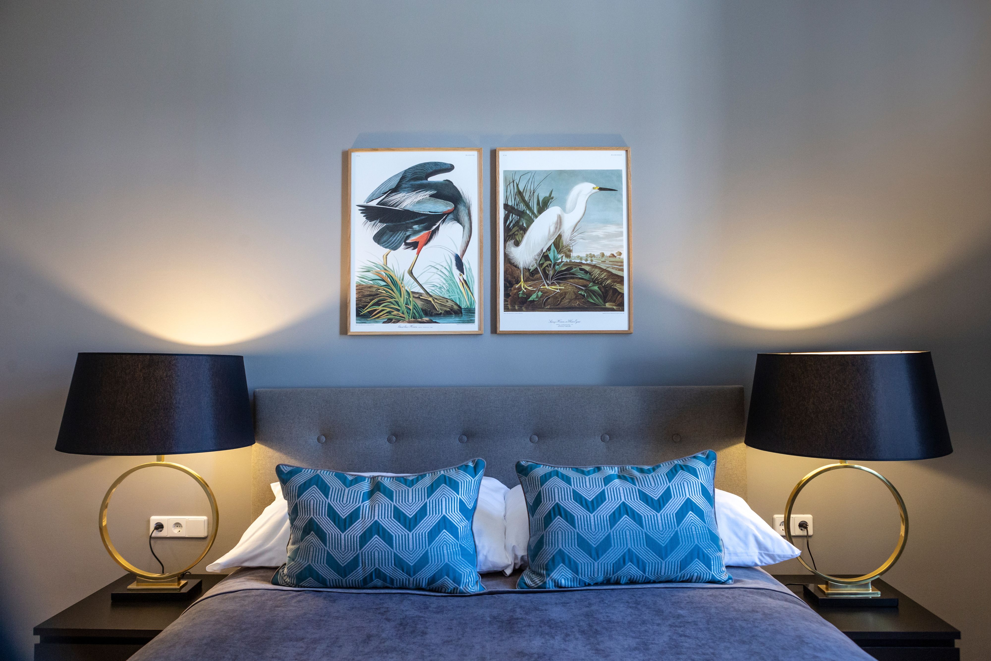 Double room with gray bed, blue pillows, two bedside lamps and bird pictures on the wall.