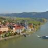 Aerial view of a village on a river with a ship, surrounded by hills and vineyards.