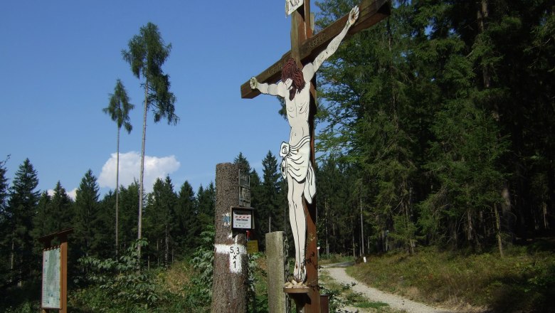 A wooden crucifix at the edge of the forest with a path in the background.