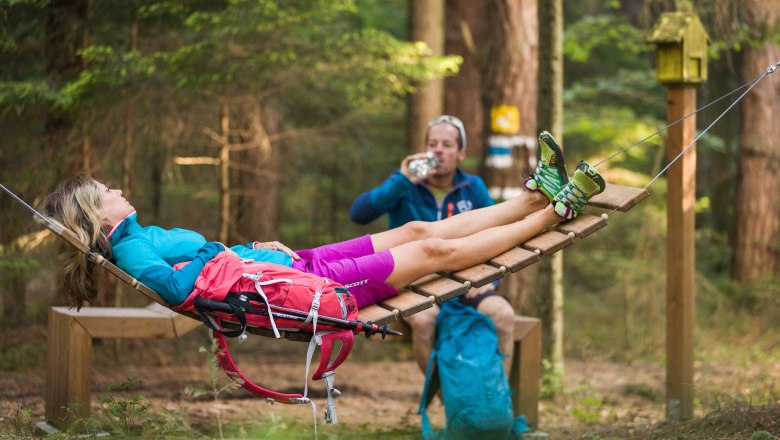 Two people relaxing in the forest, one lying in a hammock, the other drinking from a bottle.