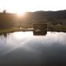 A pond with a wooden building on the shore, surrounded by hills and trees at sunset.