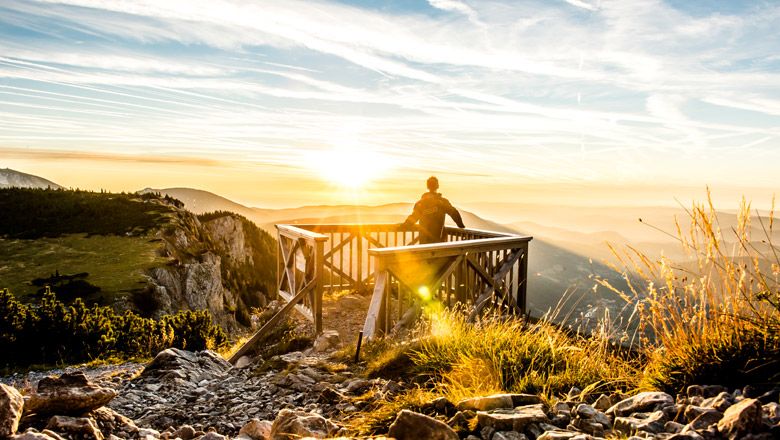 Person stands on a wooden platform and gazes into the sunset over a mountain landscape.
