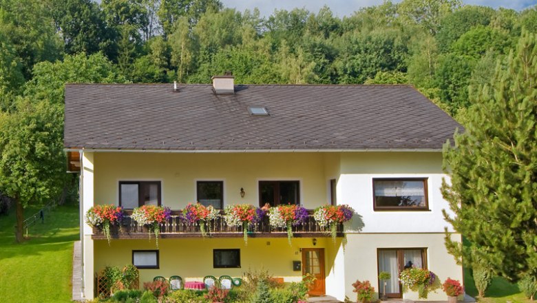 Two-storey yellow house with balcony and flowers, surrounded by trees and garden.