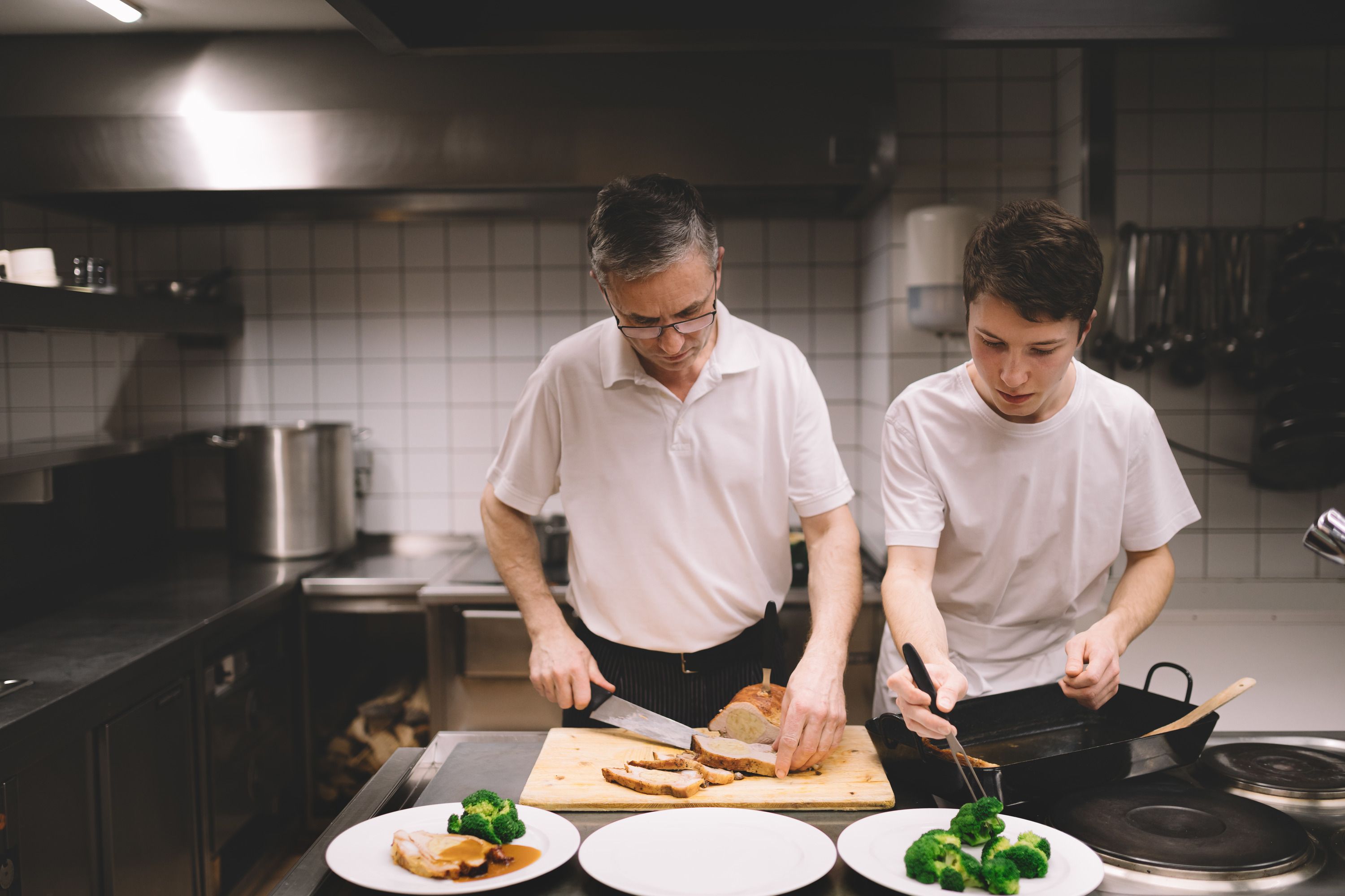 An older man and a younger man work together in a kitchen. The older man cuts meat while the younger man puts broccoli on a plate.