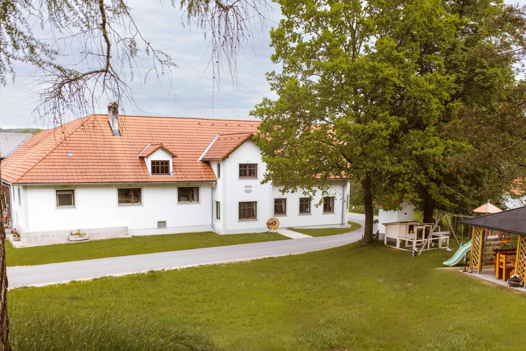 A white building with a red tiled roof, surrounded by trees and a meadow, with a playground in the foreground.