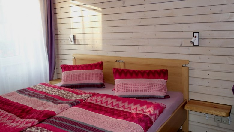 A cozy bedroom with a double bed covered with red-patterned comforters and pillows. The wall is made of light-colored wood.