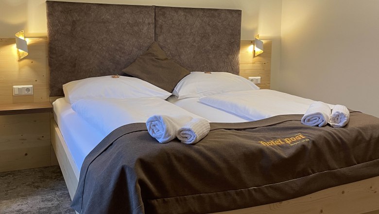 A cozy double room with a large bed, decorated with brown bed linen and towels. There are geometric patterns on the wall.