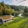 Aerial view of a modern house with a green roof, surrounded by forest and a lake.