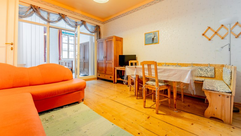 A living room with an orange sofa, wooden table and chairs, wooden floor and a cupboard. A picture hangs on the wall.