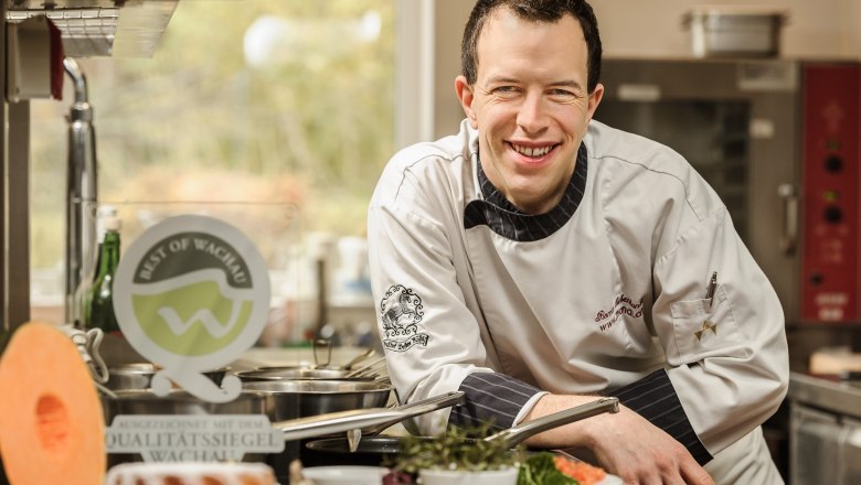 A chef in a white uniform smiles in a kitchen surrounded by fresh ingredients and a Wachau quality seal.
