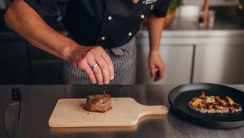 A cook seasons a piece of meat on a wooden board in a kitchen.