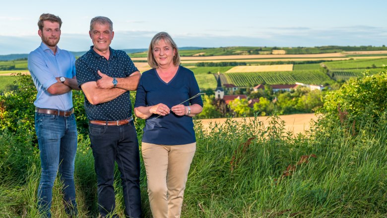 Three people are standing in a vineyard with a view of a rural landscape in the background.
