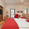 Hotel room with double bed, red bed linen, sofa, red armchair and large window.