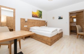 Modern bedroom with wooden furniture, double bed, table and chair. A mirror and pictures on the wall, door leading to a bathroom.