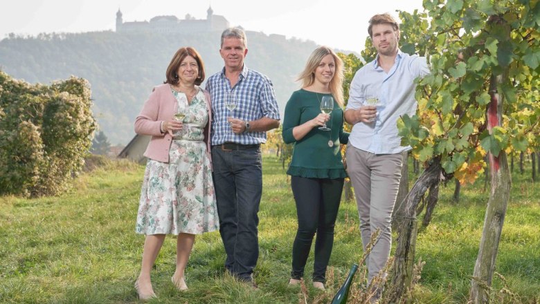 Four people are standing in a vineyard with wine glasses in their hands; a castle on a hill can be seen in the background.
